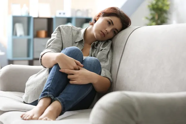 Depressão Mulher sentada no sofá abraçada as pernas com expressão triste